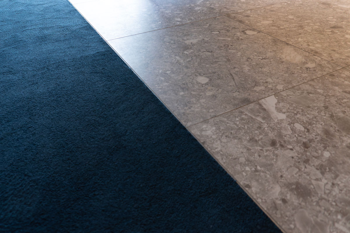 Übergang von blauem Hochflorteppich zu grauen Feinsteinzeugfliesen in Terrazzo-Optik in der KSC Premium Lounge
