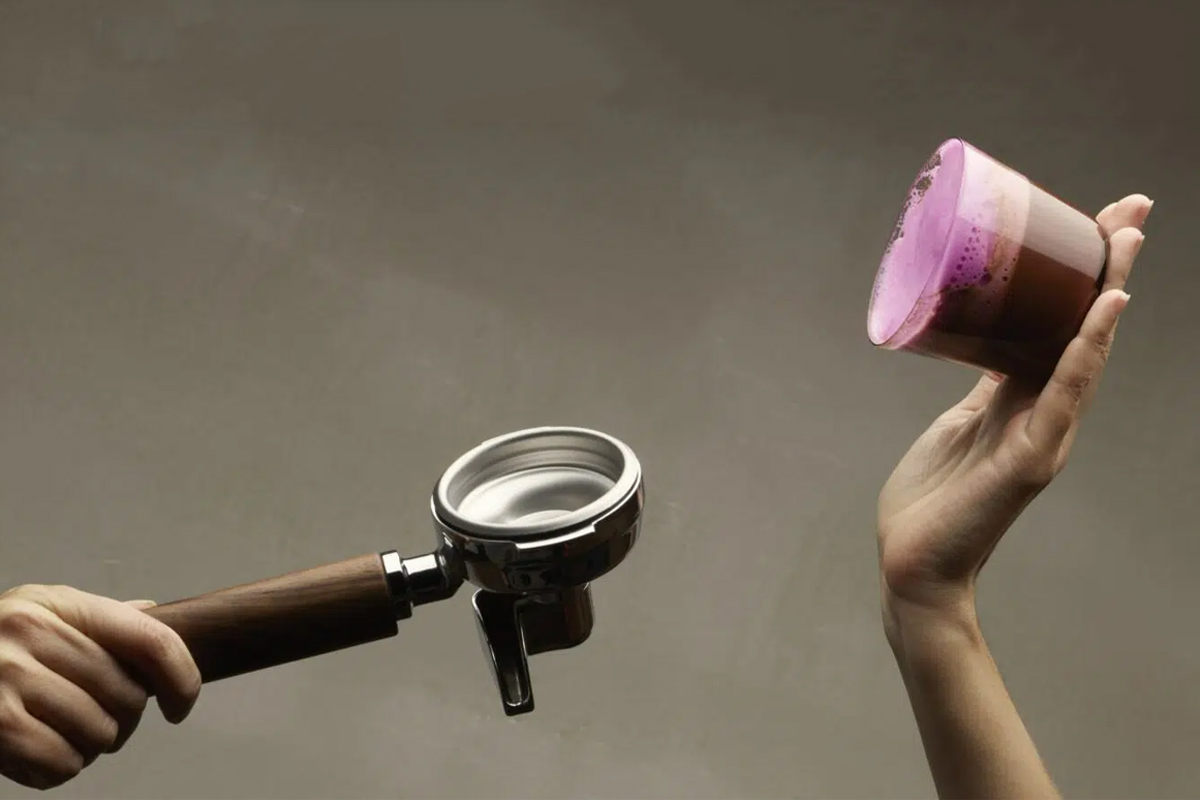 Espressokunst-LIGRE-Espresso-Maschine Zwei Hände. Eine hält den Doppel-Siebträger, eine ein Glas Kaffee mit pinkem Schaum.